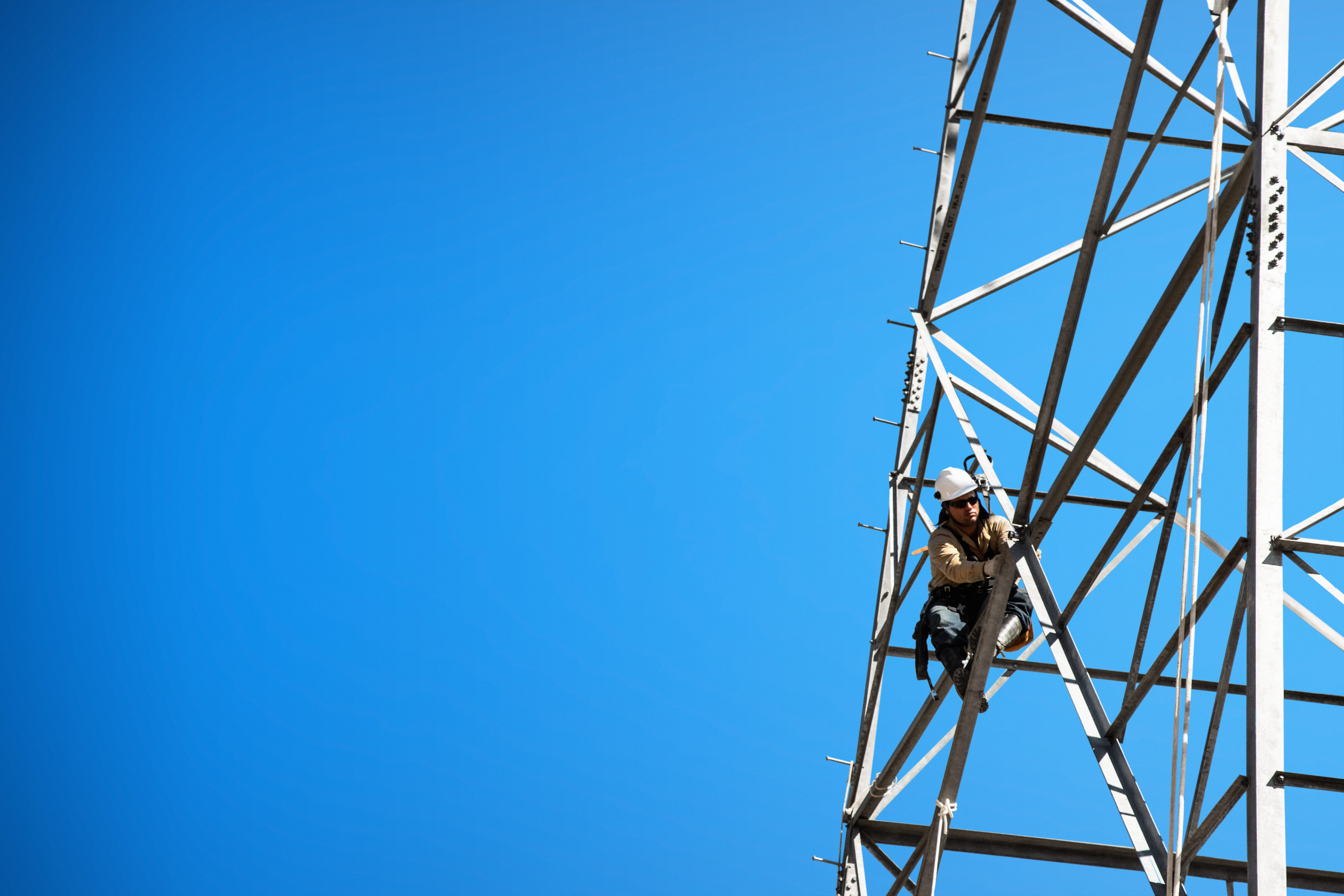 Trabalhador com equipamentos de segurança escala uma torre de metal contra um céu azul claro.