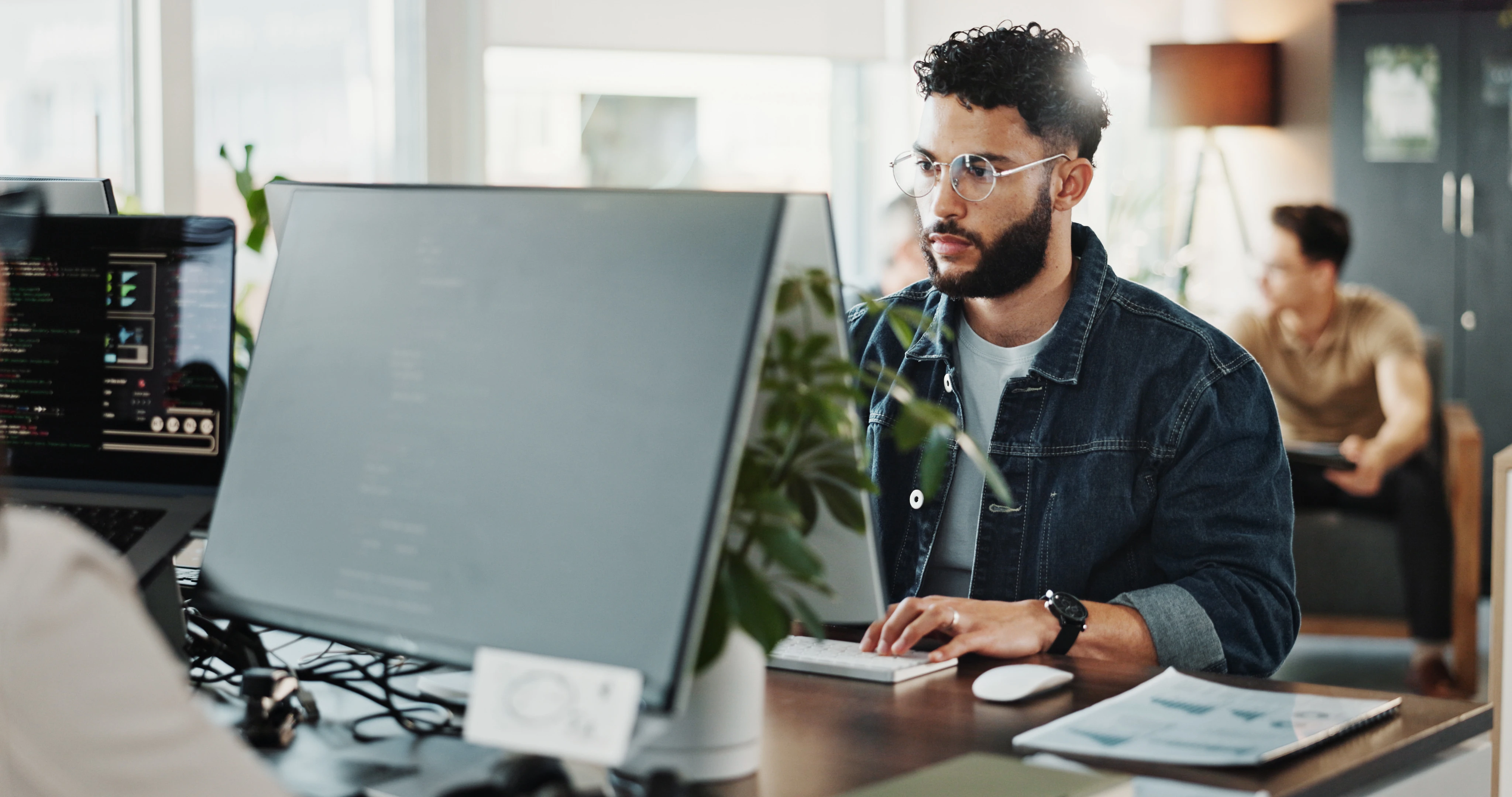 Homem pardo com barba usa óculos e está trabalhando no computador