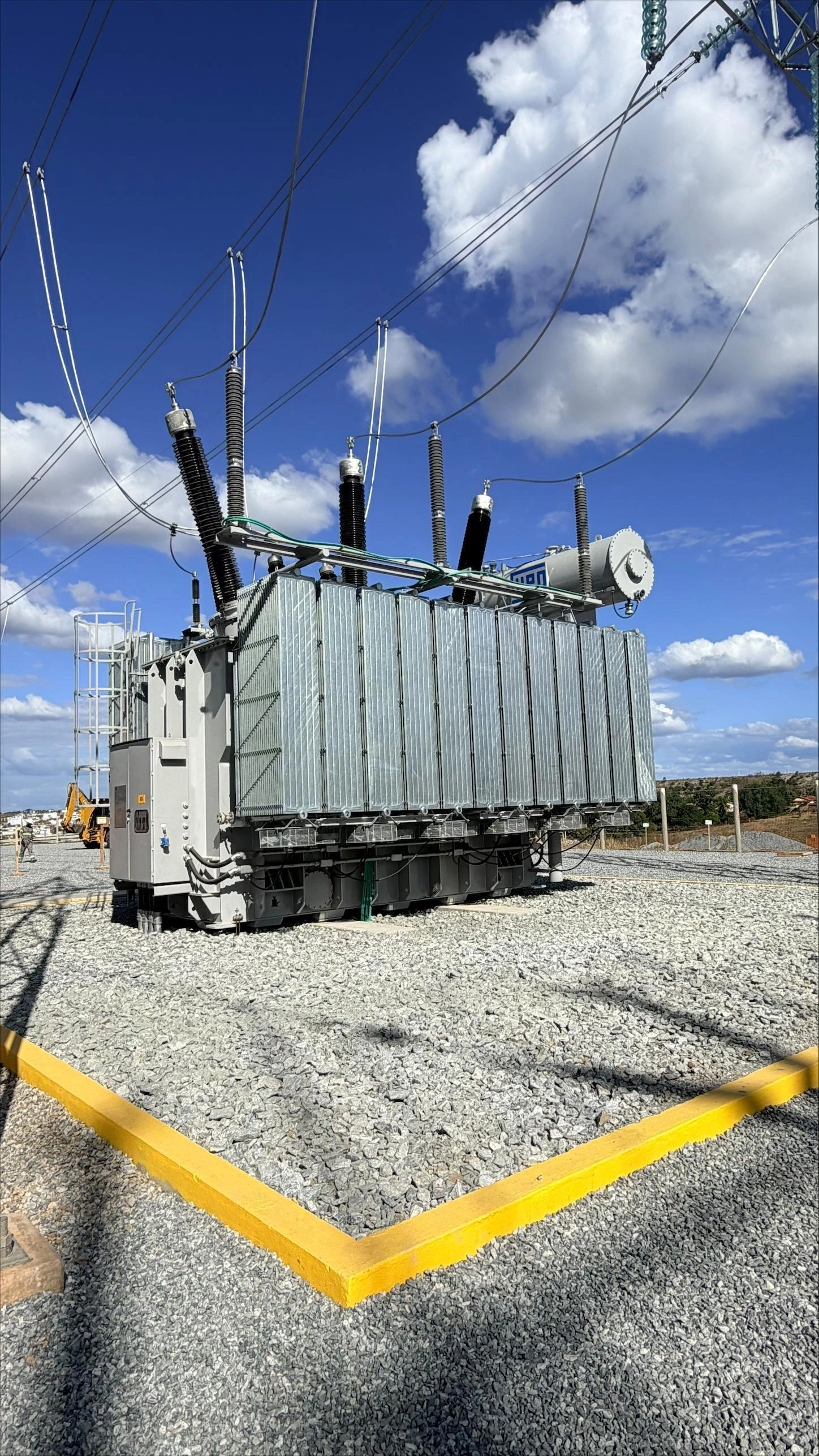 Transformador de potência instalado em subestação de energia, conectado a cabos de alta tensão, sobre piso de brita e sob céu azul com nuvens.