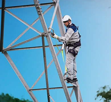 essoa utilizando equipamentos de segurança realiza atividade em uma estrutura metálica de transmissão de energia, posicionada em uma escada, com céu azul ao fundo.