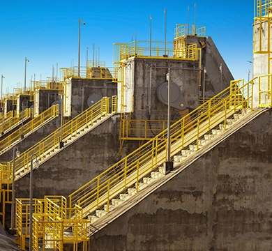 Estrutura de barragem com escadarias e passarelas metálicas amarelas, equipamentos de monitoramento e plataformas de acesso distribuídas ao longo do paredão de concreto sob céu azul.