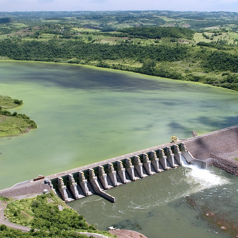 Vista aérea de uma barragem hidrelétrica com vertedouro em operação, reservatório de água ao fundo e áreas verdes ao redor.