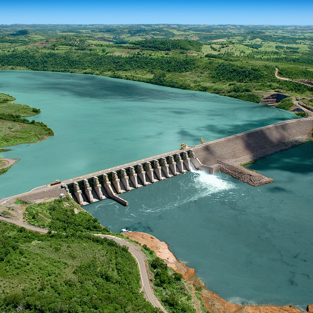 Vista aérea de uma barragem com vertedouro e reservatório de águas azul-esverdeadas, cercada por áreas de vegetação e estradas de acesso