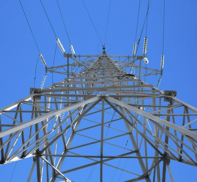 Torre de transmissão de energia vista de baixo para cima, com cabos e isoladores em destaque contra o céu azul.