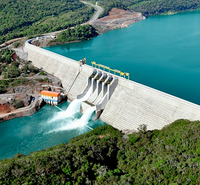 Vista aérea de uma barragem hidrelétrica com comportas abertas, liberando água para o rio, cercada por reservatório de água azul e áreas de vegetação.