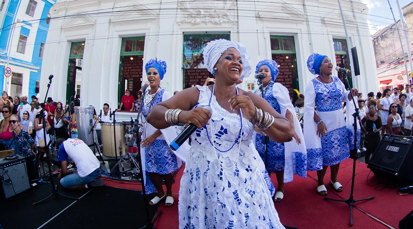 Grupo de mulheres em trajes tradicionais brancos e azuis se apresenta cantando e dançando em uma rua movimentada, diante de um prédio histórico, durante uma celebração cultural.