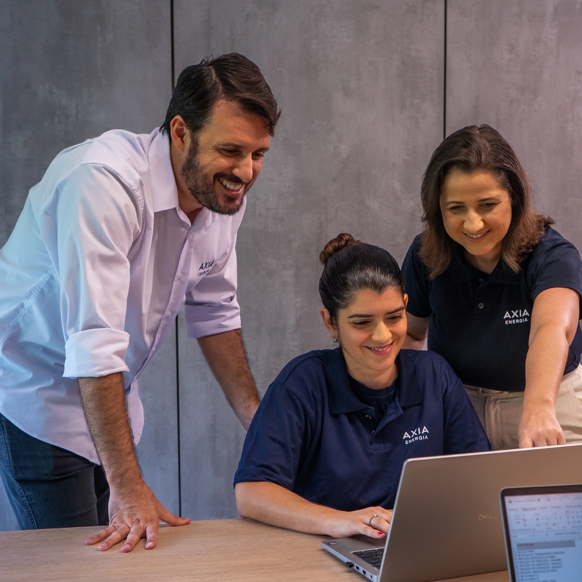 Profissionais da AXIA Energia reunidos em frente a um notebook, analisando informações em ambiente corporativo.