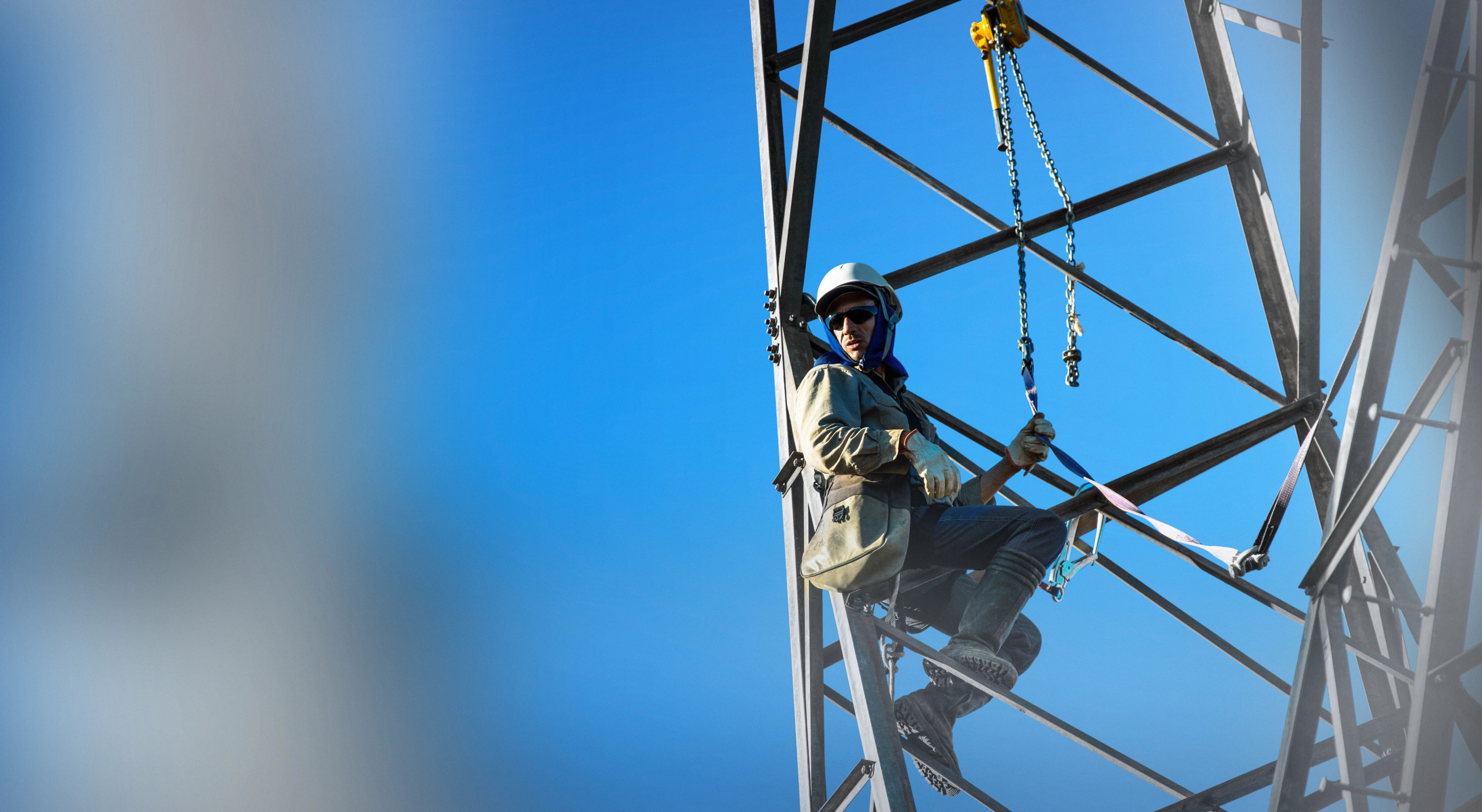 Trabalhador usando capacete e óculos de proteção está suspenso por cordas e correntes em uma estrutura metálica alta, realizando trabalho em altura contra um céu azul limpo.