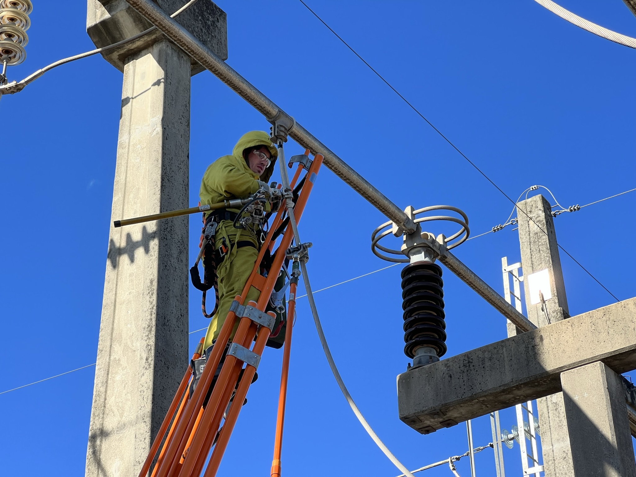 Eletricista de alta tensão, vestindo um traje de proteção amarelo completo e capacete, posicionado em uma escada laranja enquanto realiza manutenção em um barramento de uma subestação de energia, com o céu azul ao fundo.