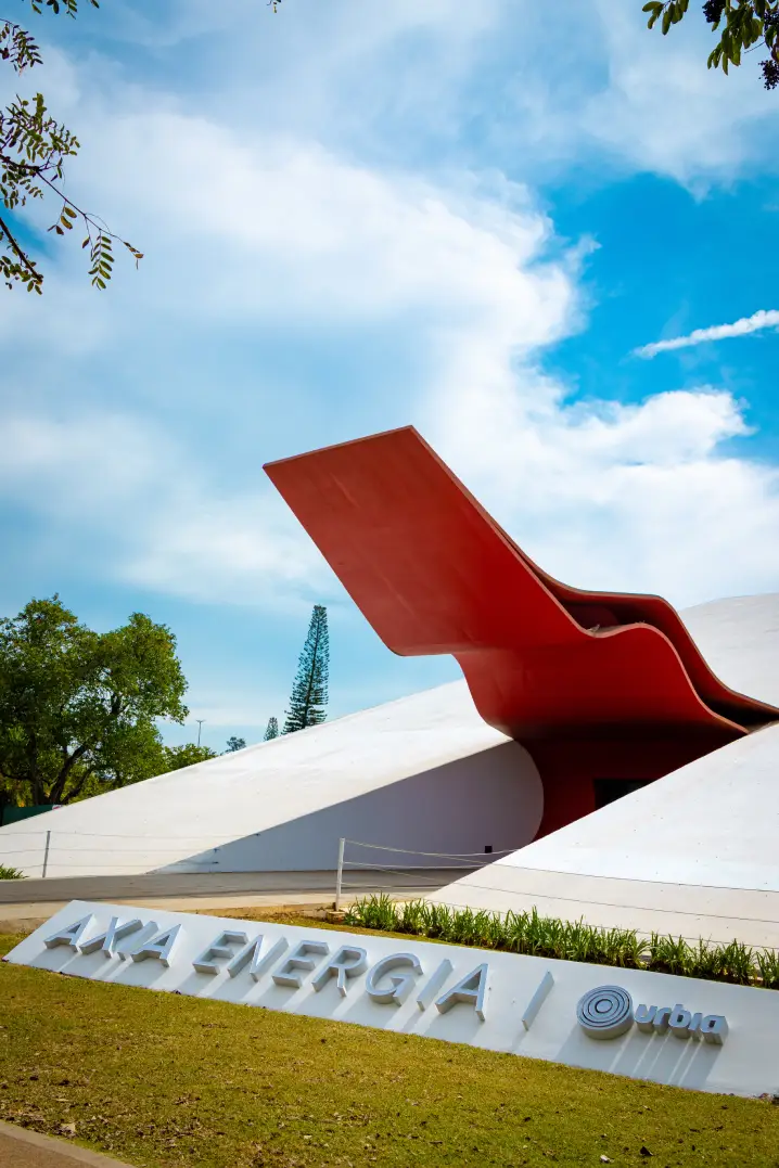 Vista externa do Auditório AXIA Ibirapuera, projetado por Oscar Niemeyer, com destaque para a marquise vermelha característica da fachada, áreas verdes ao redor e a identificação do espaço em primeiro plano.