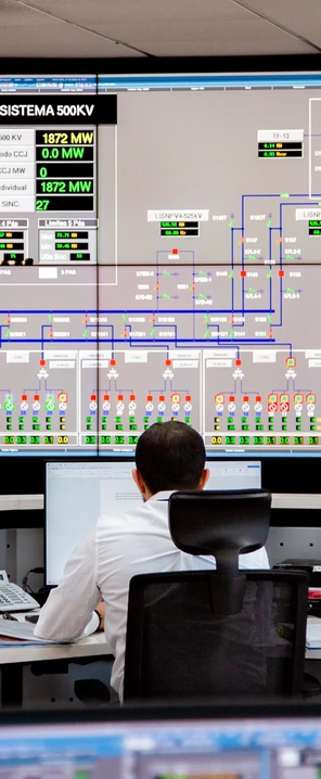 Profissional em sala de controle, diante de monitores e de um grande painel digital com diagramas e indicadores de um sistema elétrico.