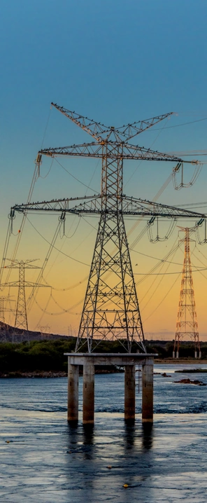 Torres e linhas de transmissão de energia atravessam uma paisagem com rio e vegetação, sob céu azul e dourado ao entardecer.