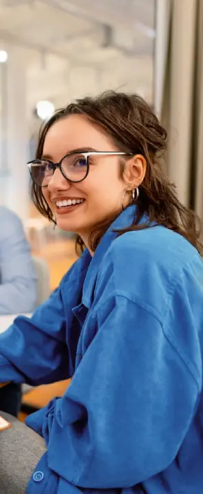 Mulher jovem branca de óculos interagindo com um homem de óculos, enquanto duas pessoas aparecem desfocadas ao fundo