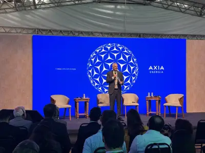 Pessoa palestrando em palco no Energy Summit, com público à frente e fundo azul com o logo da AXIA Energia.