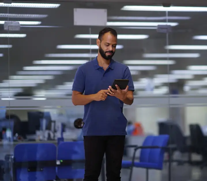 A person stands in a modern office environment, holding a tablet with both hands while interacting with the screen. In the background, there are workstations and typical corporate lighting.