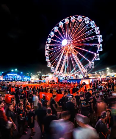 Roda-gigante iluminada durante evento noturno, cercada por grande público e estruturas de entretenimento em área aberta.