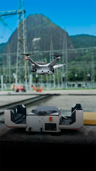 Drone sobrevoa uma área de subestação elétrica, com estação de carregamento em primeiro plano e estruturas de transmissão ao fundo, tendo uma formação rochosa ao longe sob céu azul.