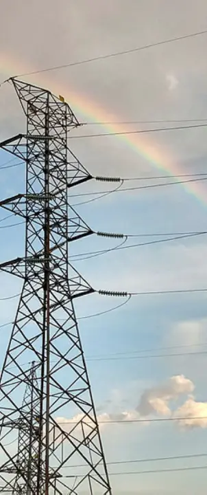 Torre de transmissão de energia elétrica com estrutura metálica e cabos de alta tensão em destaque, vista contra um céu parcialmente nublado. Ao fundo, um arco-íris cruza a paisagem, criando contraste entre a infraestrutura elétrica e os elementos naturais.