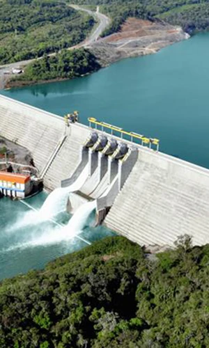 Imagem aérea de uma barragem de concreto sobre um rio, com água sendo liberada por comportas centrais. À direita, um grande reservatório de água azul; ao redor, áreas verdes e vias de acesso próximas à estrutura.